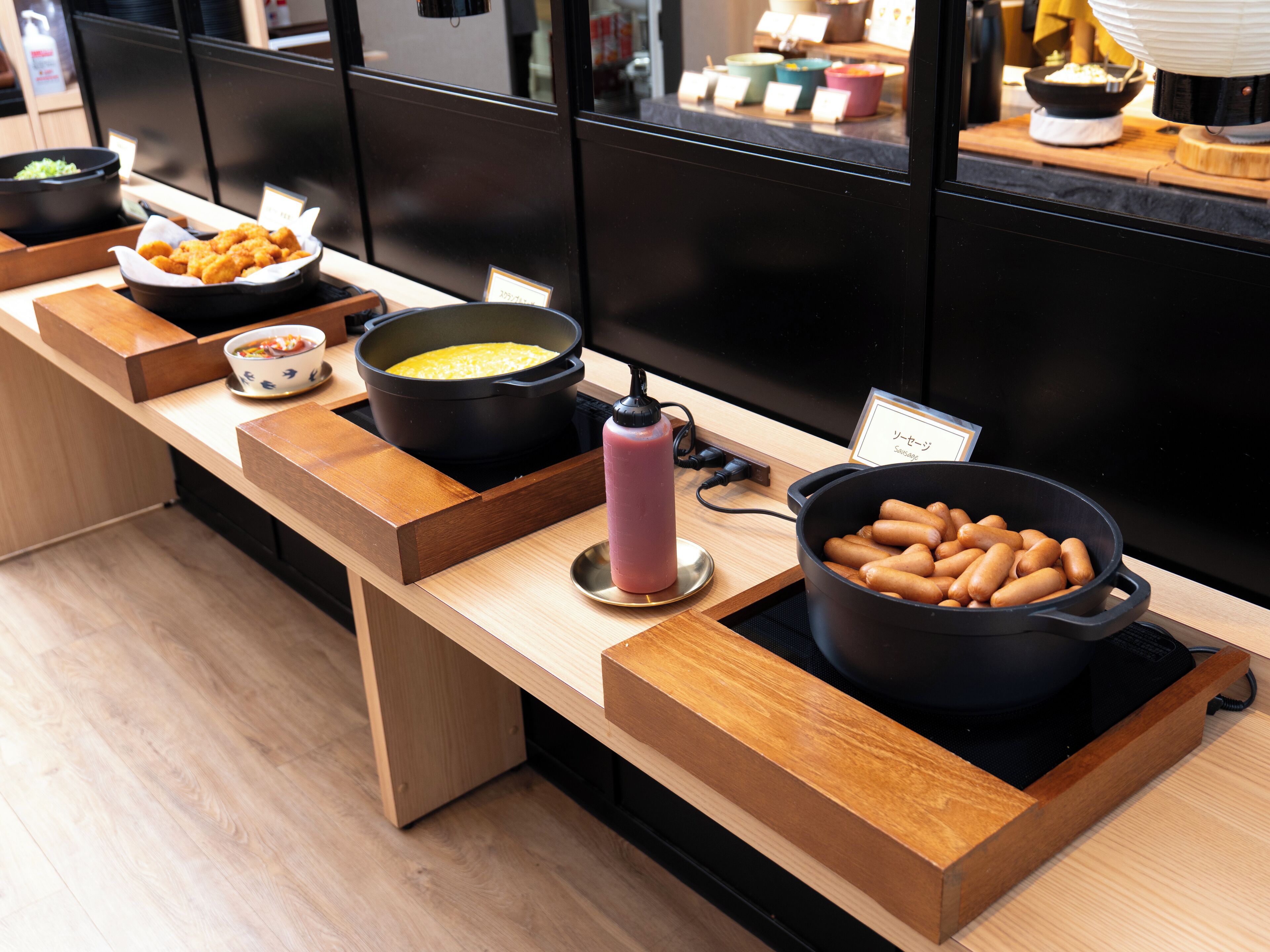 A buffet setup with various dishes including a pot of sausages, a bowl of soup, and a container of fried items.