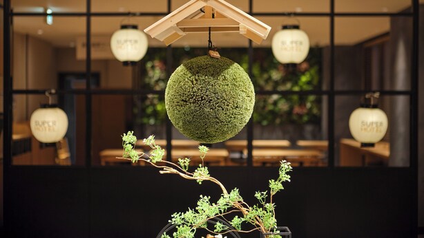 A hanging spherical decoration with a wooden hanger, a potted plant with green foliage, and multiple hanging light fixtures with the text "SUPER HOTEL" in the background. - Super Hotel Kyoto Shijyo Kawaramachi (Kyoto)