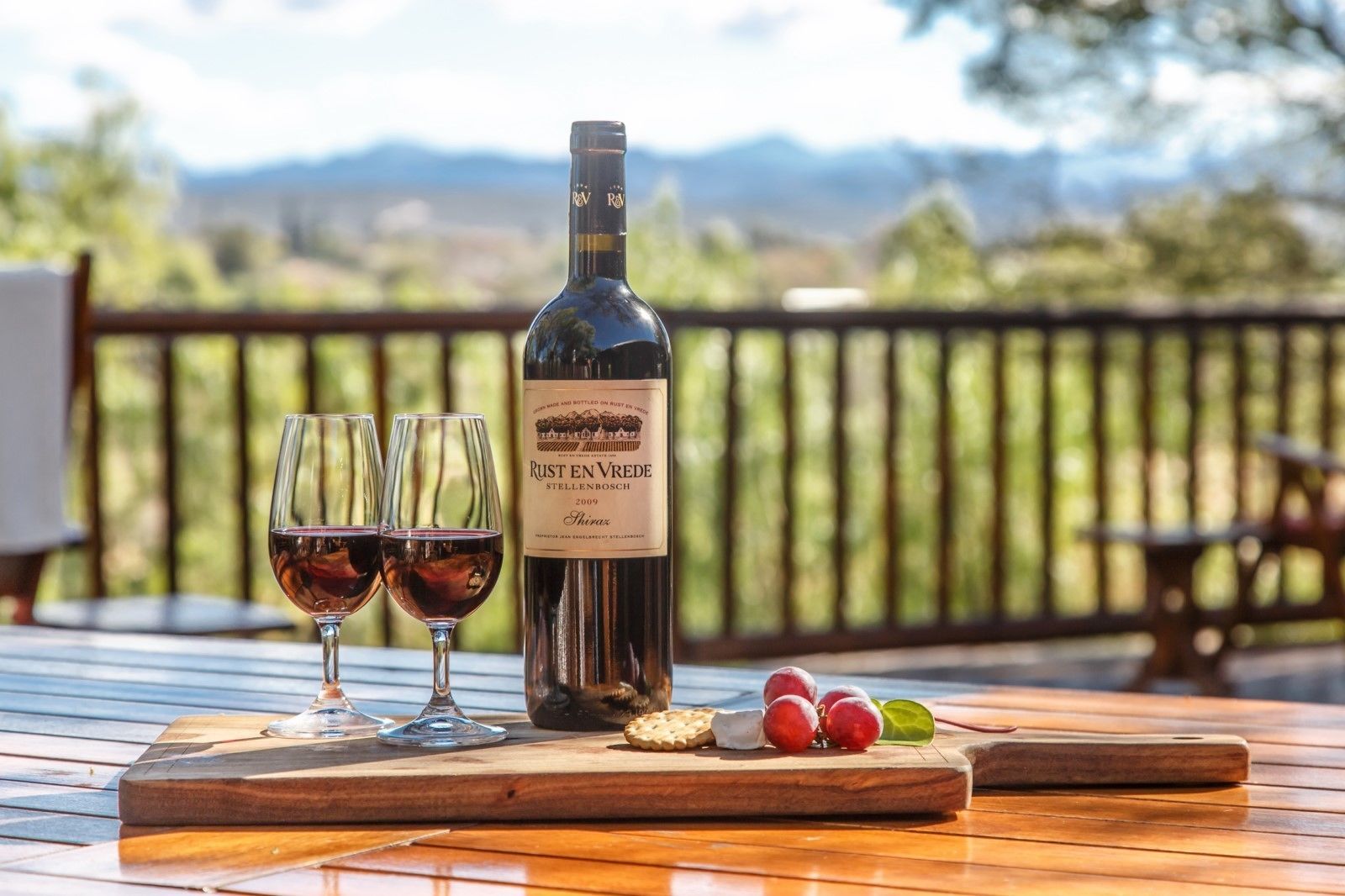 Eine Flasche Rust en Vrede Wein, zwei Gläser und einige Trauben auf einem Holztisch mit einer schönen Aussicht im Hintergrund.