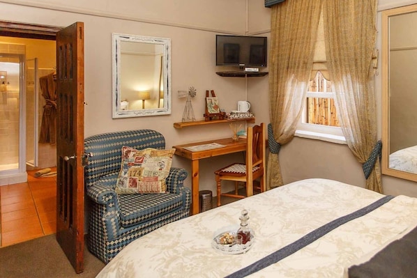 A bedroom with a bed, a chair, a desk, a television, and a window with curtains. - Riverside Lodge (Oudtshoorn)