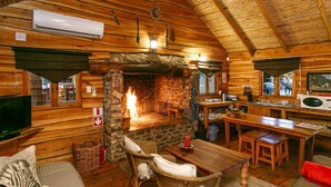 A cozy cabin interior with a stone fireplace, wooden furniture, and a kitchen area. - Riverside Lodge (Oudtshoorn)