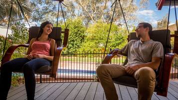 Two people sitting on a wooden swing chair with a metal fence and a pool in the background.