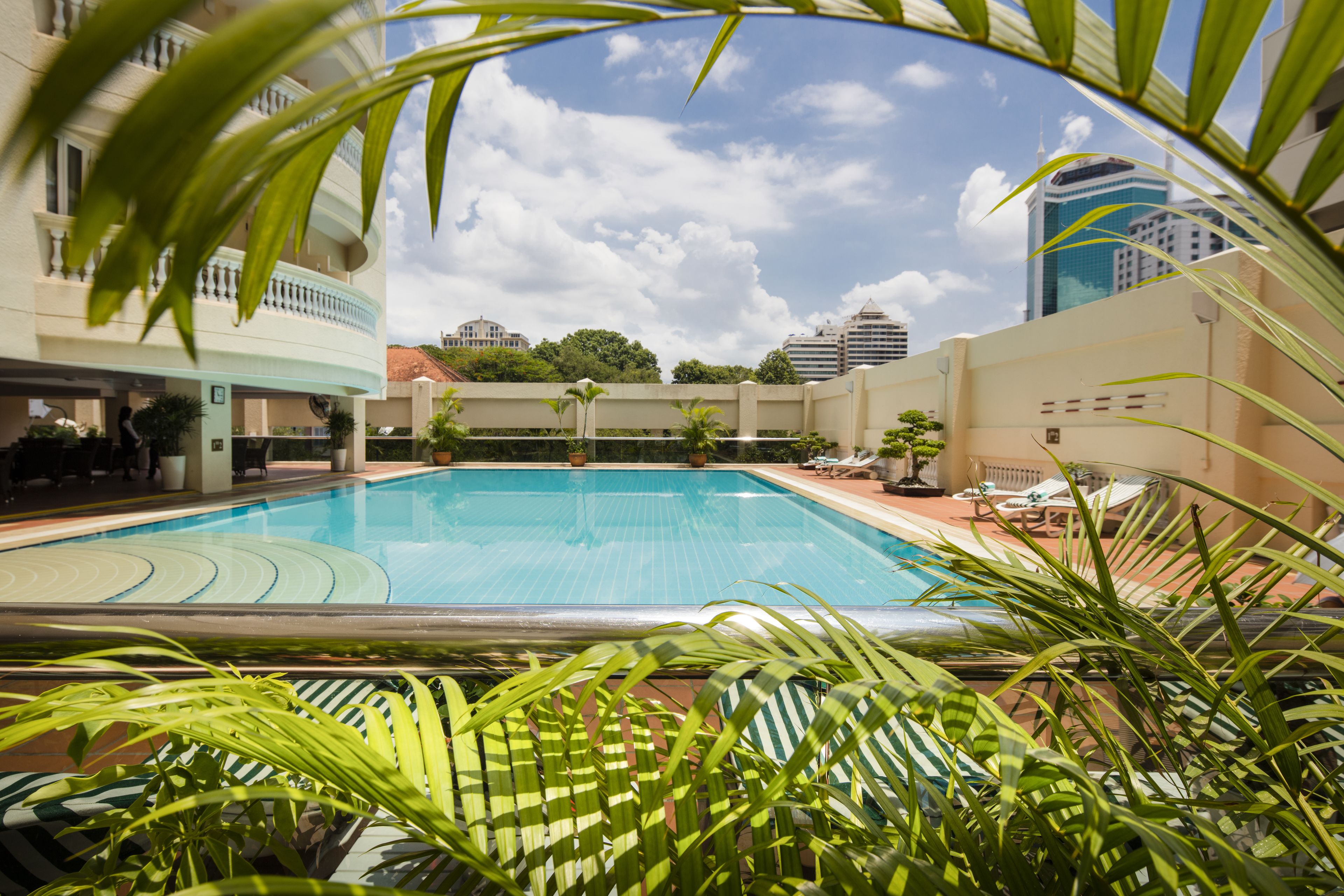 A swimming pool with a view of a cityscape, surrounded by greenery and a building with a balcony.