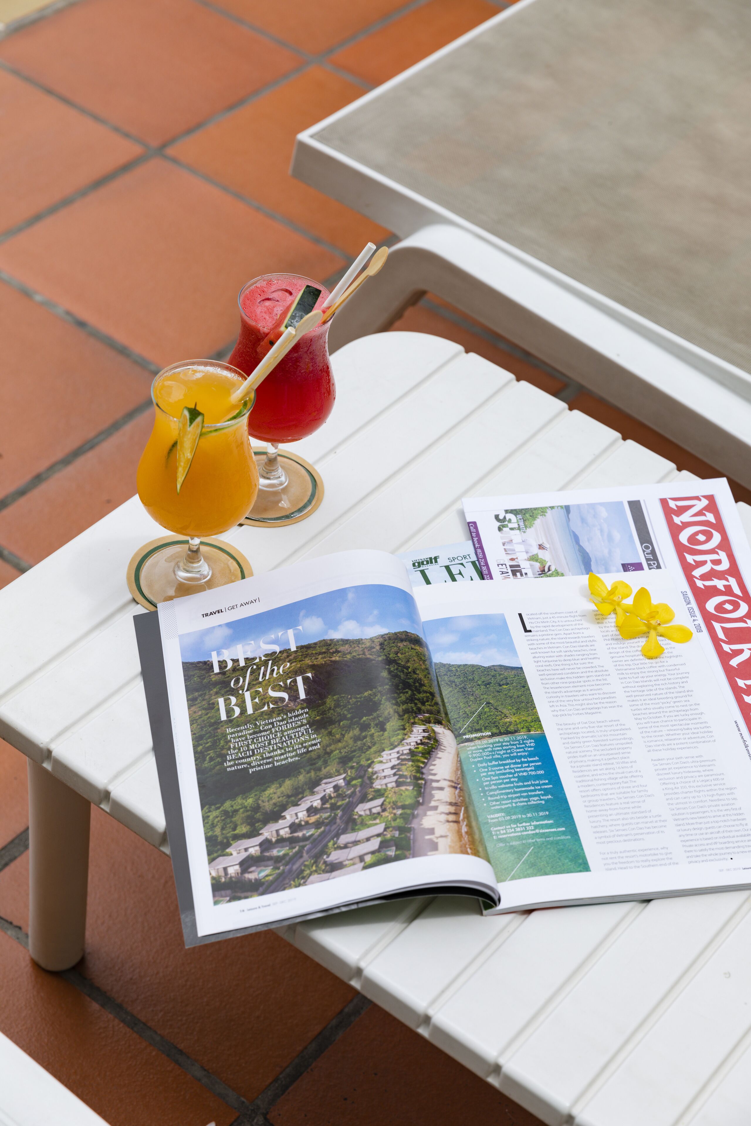 A magazine open on a white table with drinks and a flower decoration.