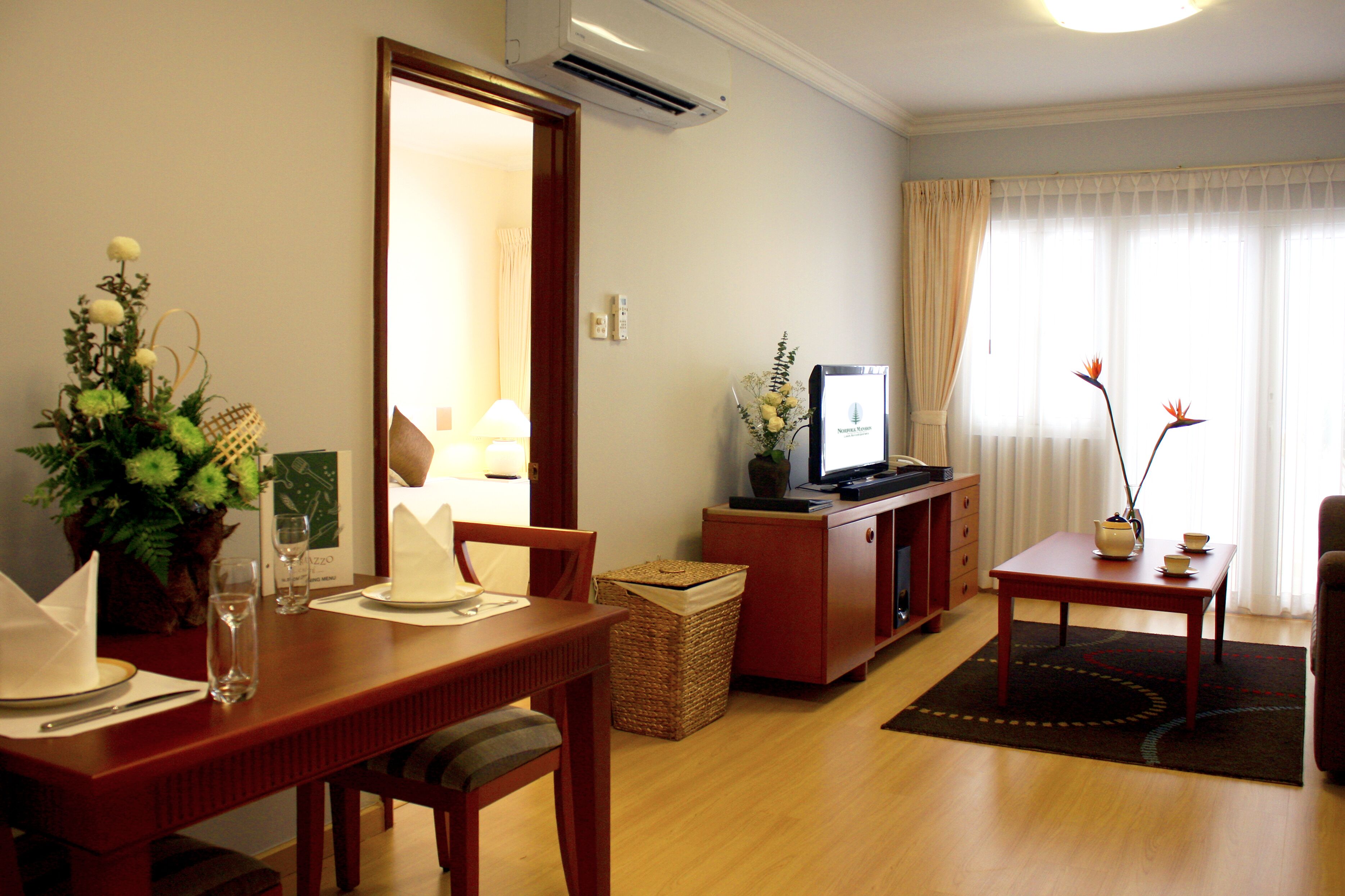 A dining area with a wooden table set for two, a television on a stand, and a window with curtains.