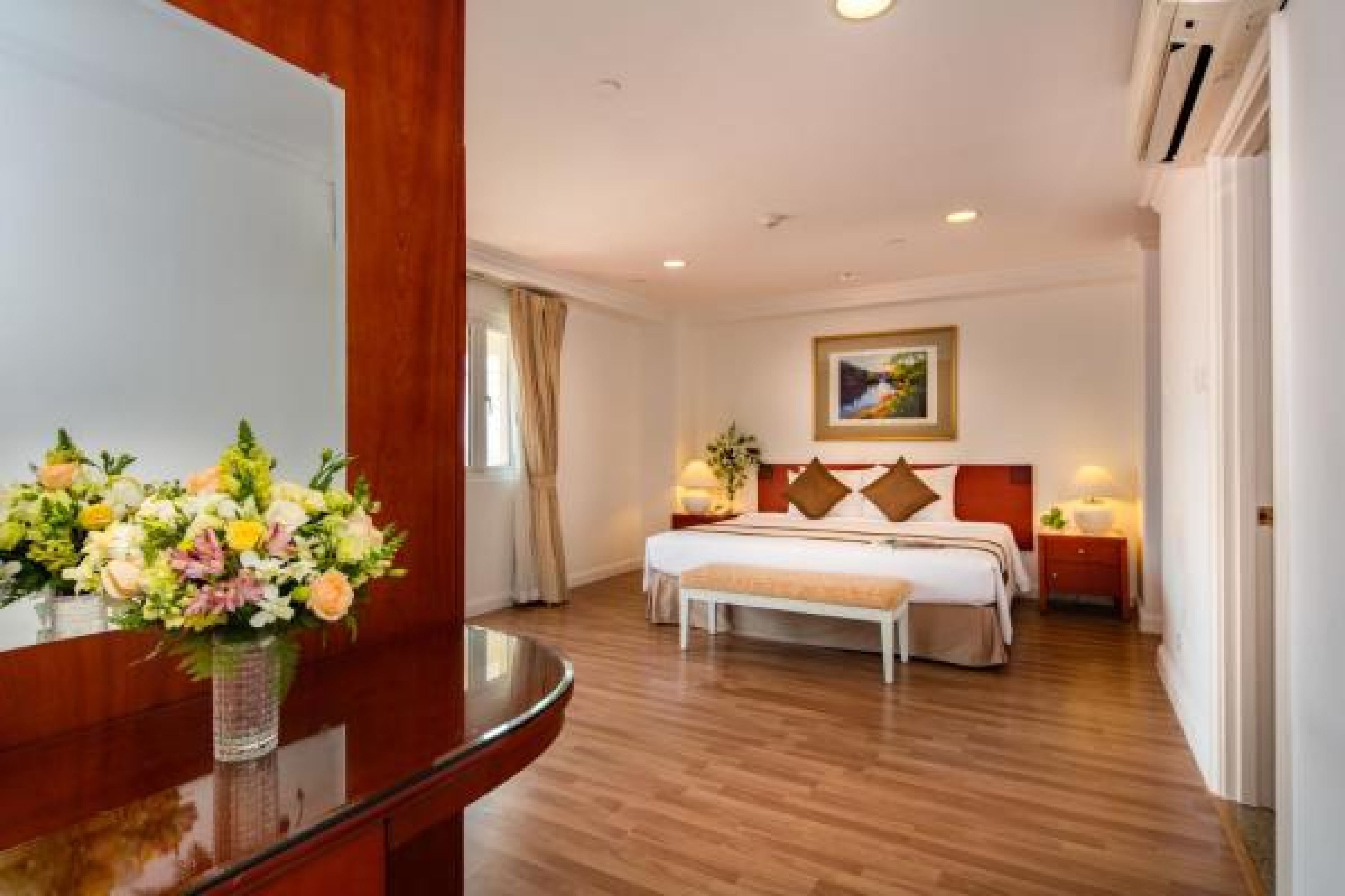 A hotel room with a wooden floor, a bed with a white headboard, a bedside table with a lamp, and a large mirror.