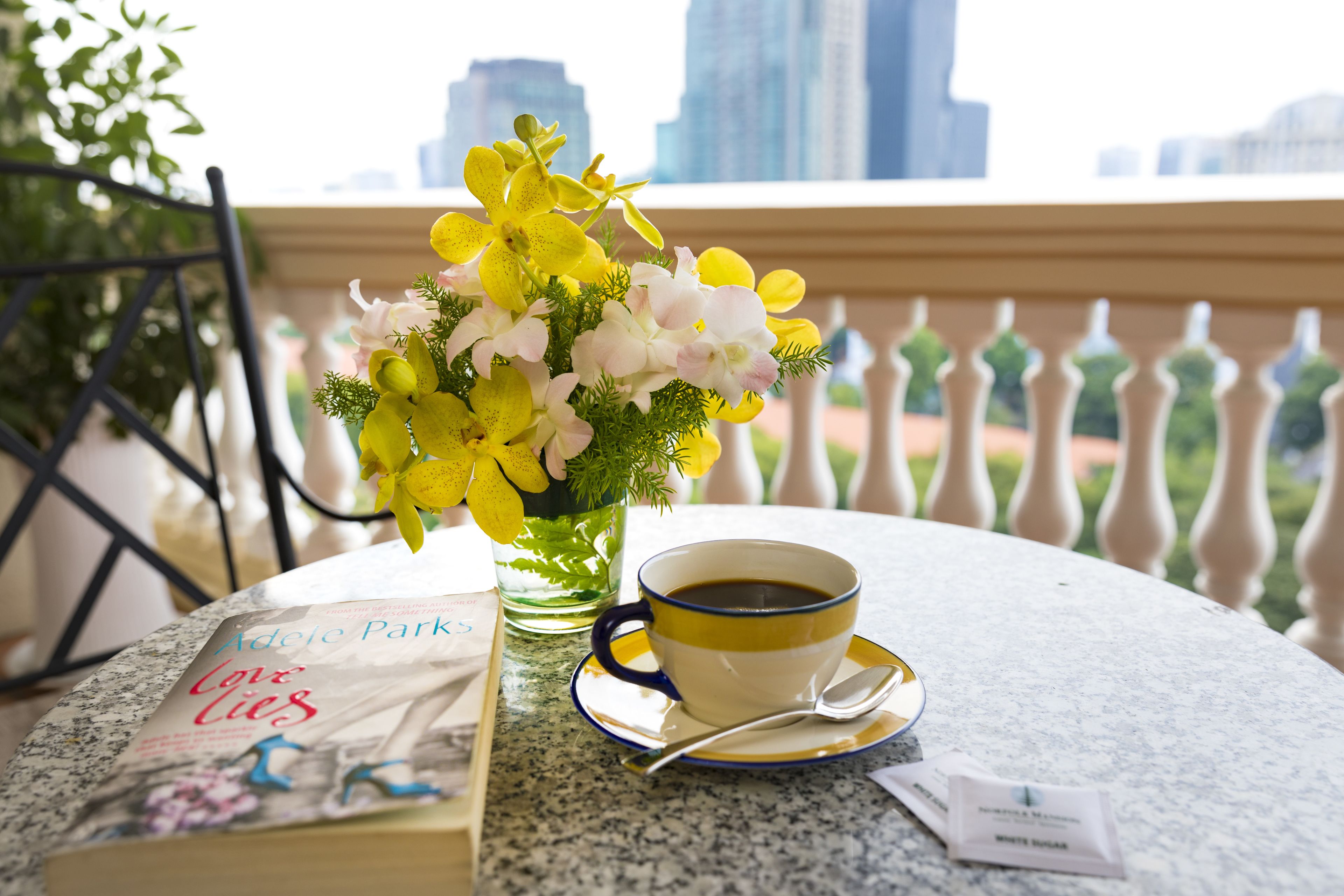 Une table sur laquelle se trouvent un livre intitulé « Les mensonges amoureux d’Adèle Park », une tasse de café et un vase rempli de fleurs jaunes.