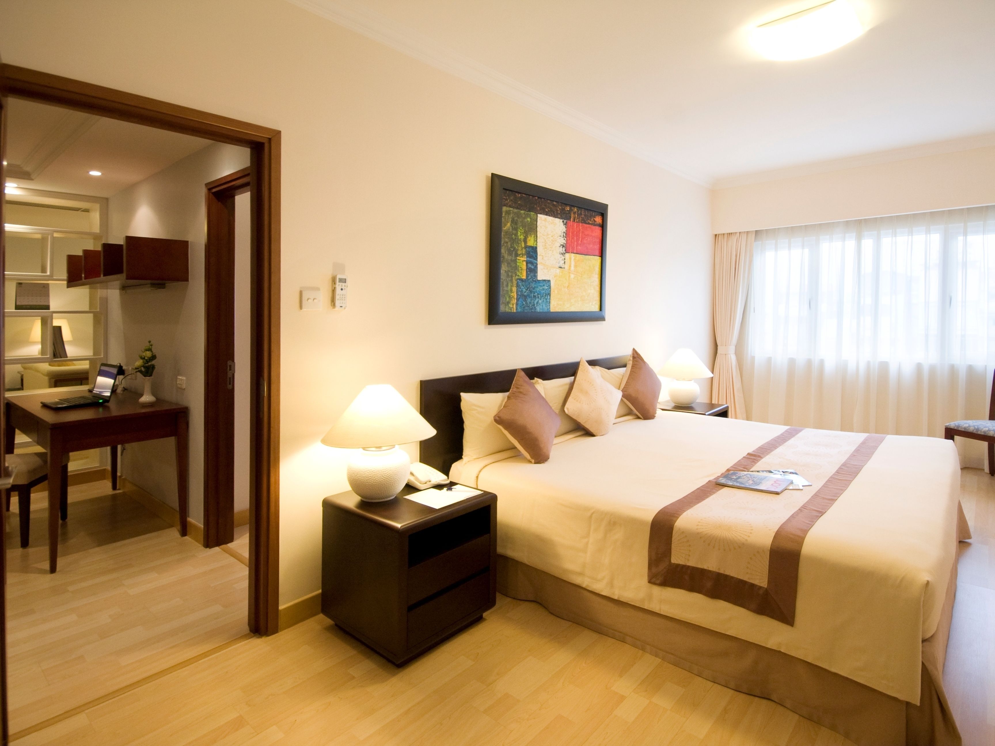 Una habitación de hotel con cama, mesita de noche, lámpara, escritorio con una computadora portátil y un cuadro en la pared.