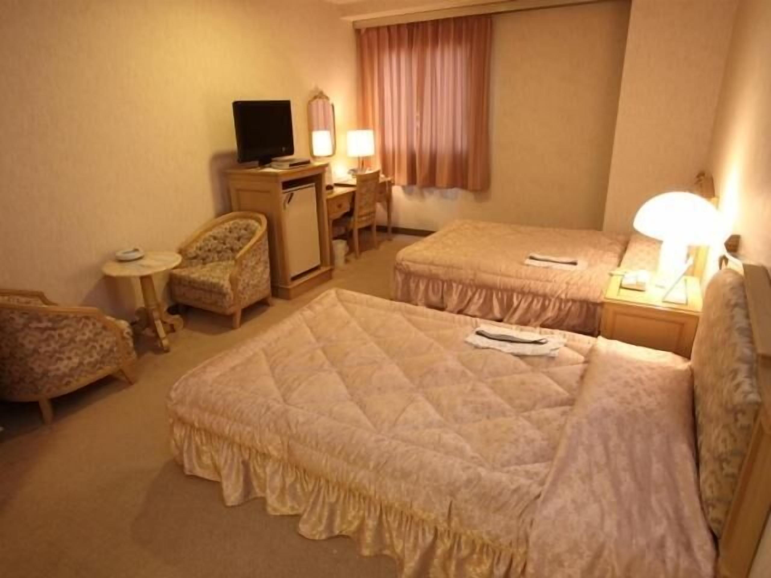 A hotel room with two beds, a desk, a chair, a television, and a window with curtains.