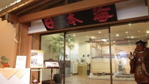 A traditional Japanese restaurant entrance with a wooden facade, a large red sign, and a statue of a figure holding a bottle. - Nogami President Hotel (Iizuka)