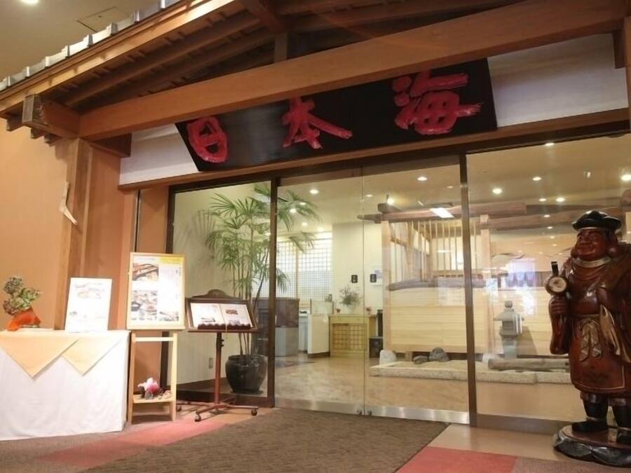 A traditional Japanese restaurant entrance with a wooden facade, a large red sign, and a statue of a figure holding a bottle.