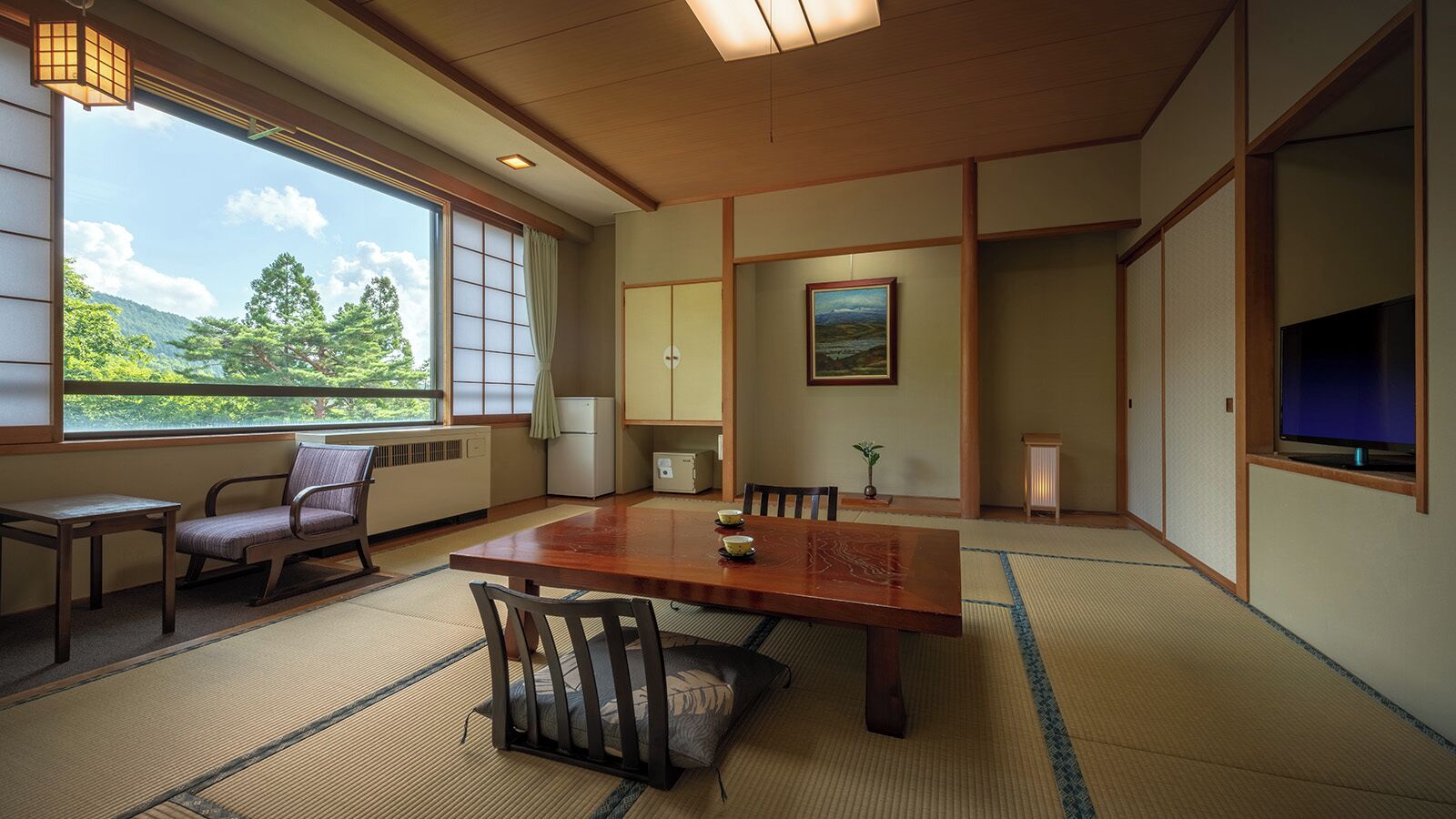 A traditional Japanese room with tatami flooring, a wooden table, chairs, a television, and a view of greenery outside.