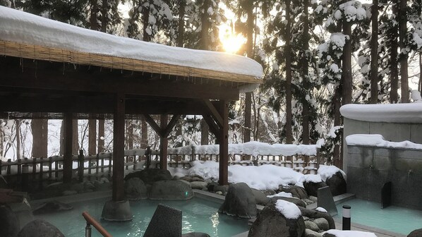 Un onsen japonés tradicional con estructura de madera, tinaja de piedra y un paisaje nevado circundante.