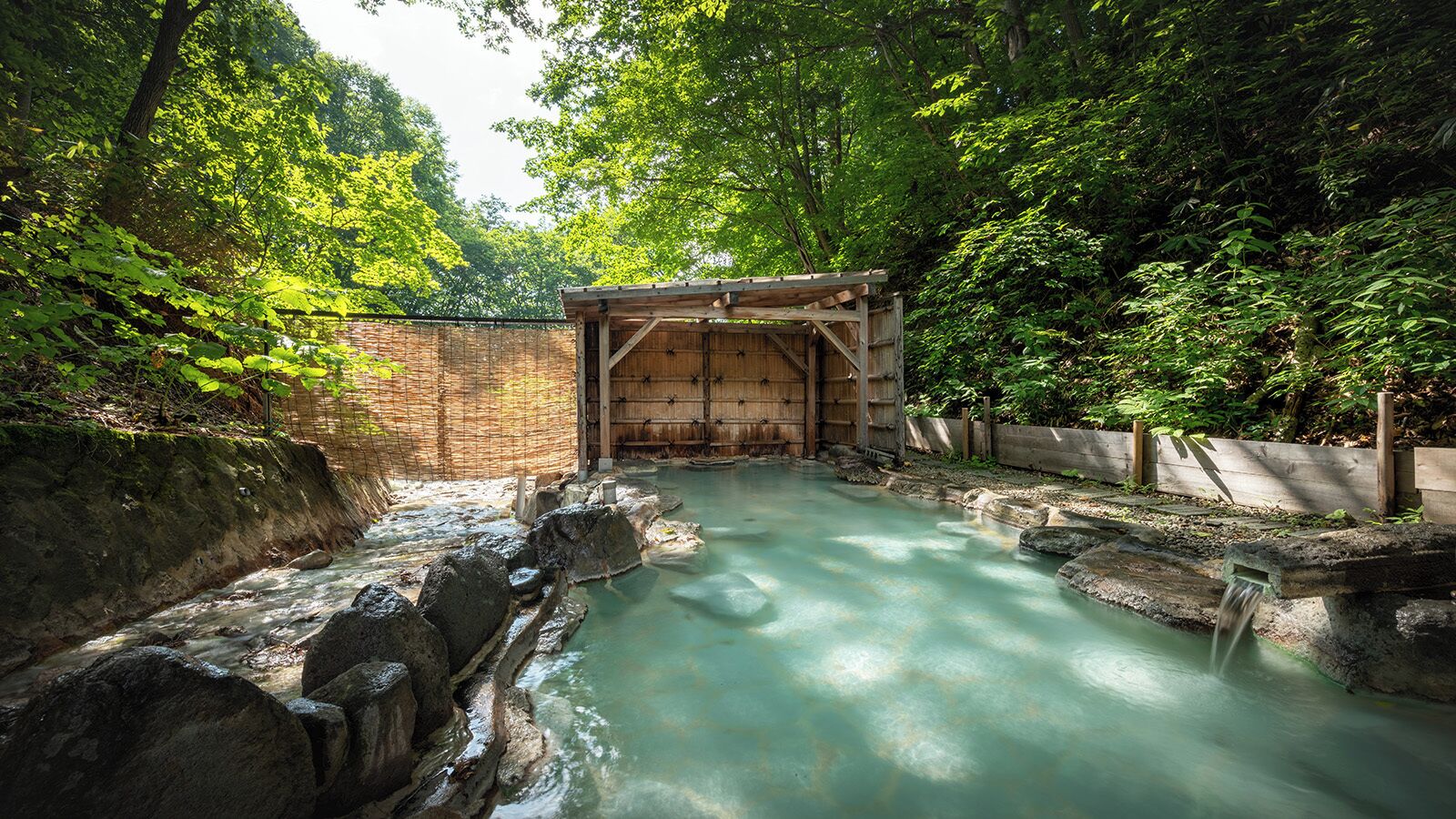 A traditional wooden bathhouse with a natural pool, surrounded by lush greenery.