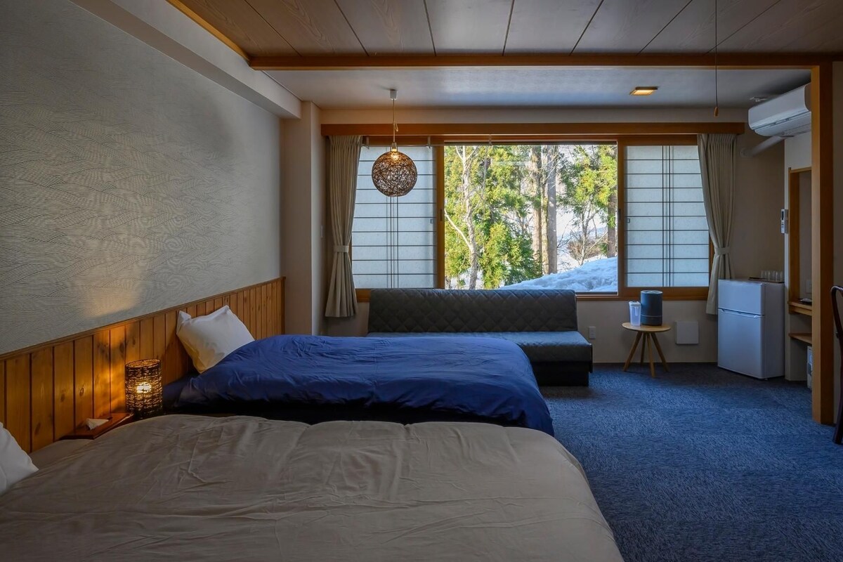 Habitación de hotel con dos camas, un escritorio, una silla, una mesita y una ventana con vistas a árboles.