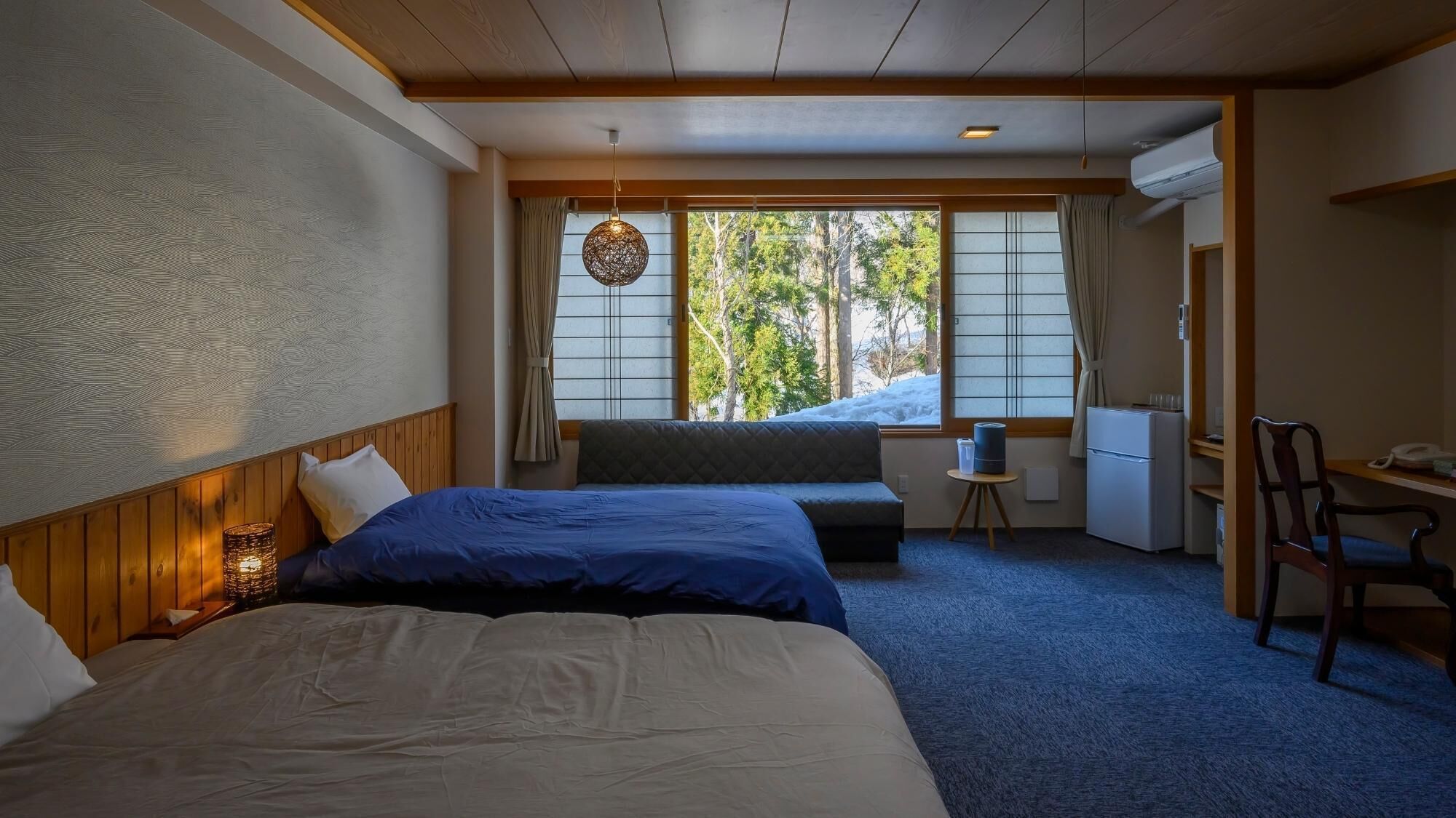 A hotel room with two beds, a desk, a chair, a small table, and a window with a view of trees.