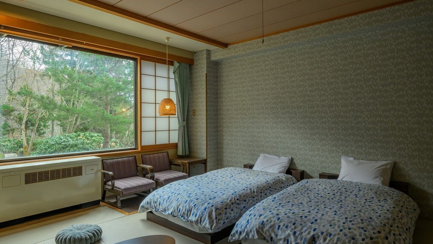 A room with two beds, a window with a view of trees, a traditional Japanese-style hanging lamp, and a radiator.