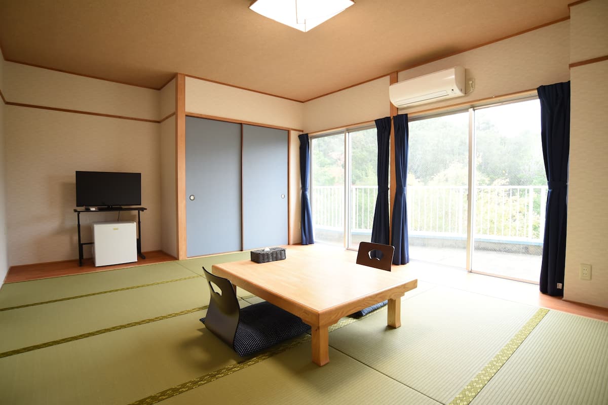 A room with tatami flooring, a wooden table, a television on a stand, and a sliding door leading to a balcony.
