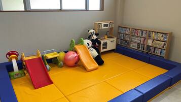 A play area with a yellow padded floor, a red slide, a small kitchen setup, and a bookshelf filled with books.