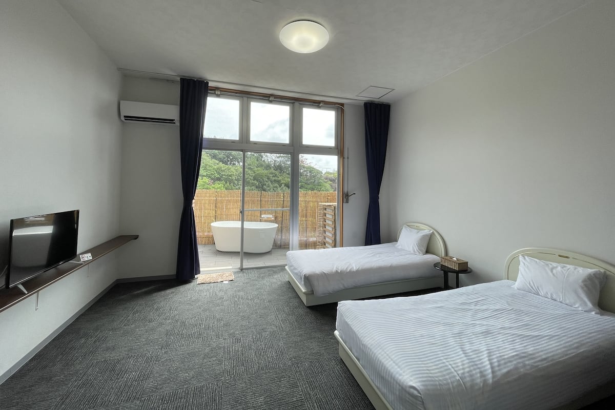 A hotel room with two beds, a TV, a small table, and a view of a garden through a large window.