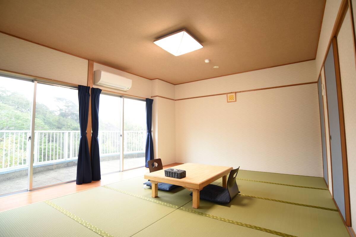 A room with tatami mat flooring, a wooden table, a chair, and a sliding glass door leading to a balcony.