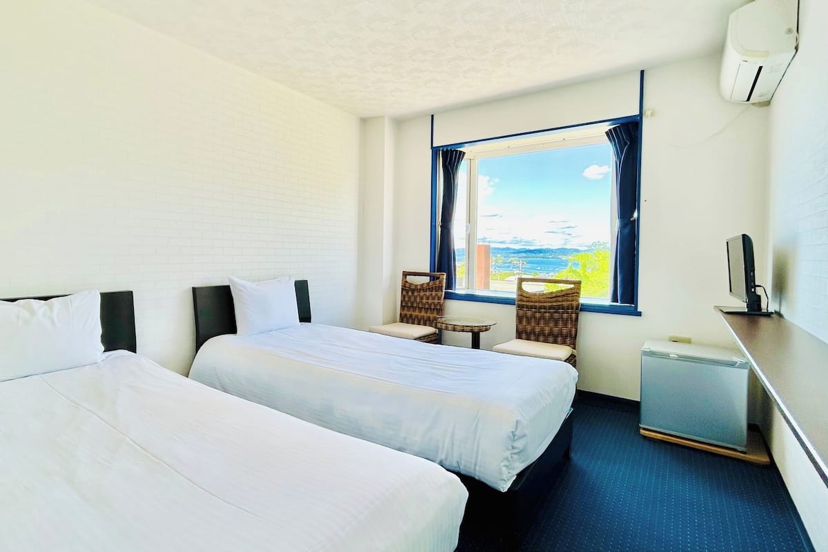 A hotel room with two beds, a small table, a chair, a TV, and a window with a view.