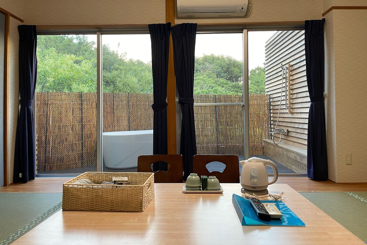 A room with a sliding glass door to a balcony, a wicker basket, a table with a kettle and cups, and a remote control.