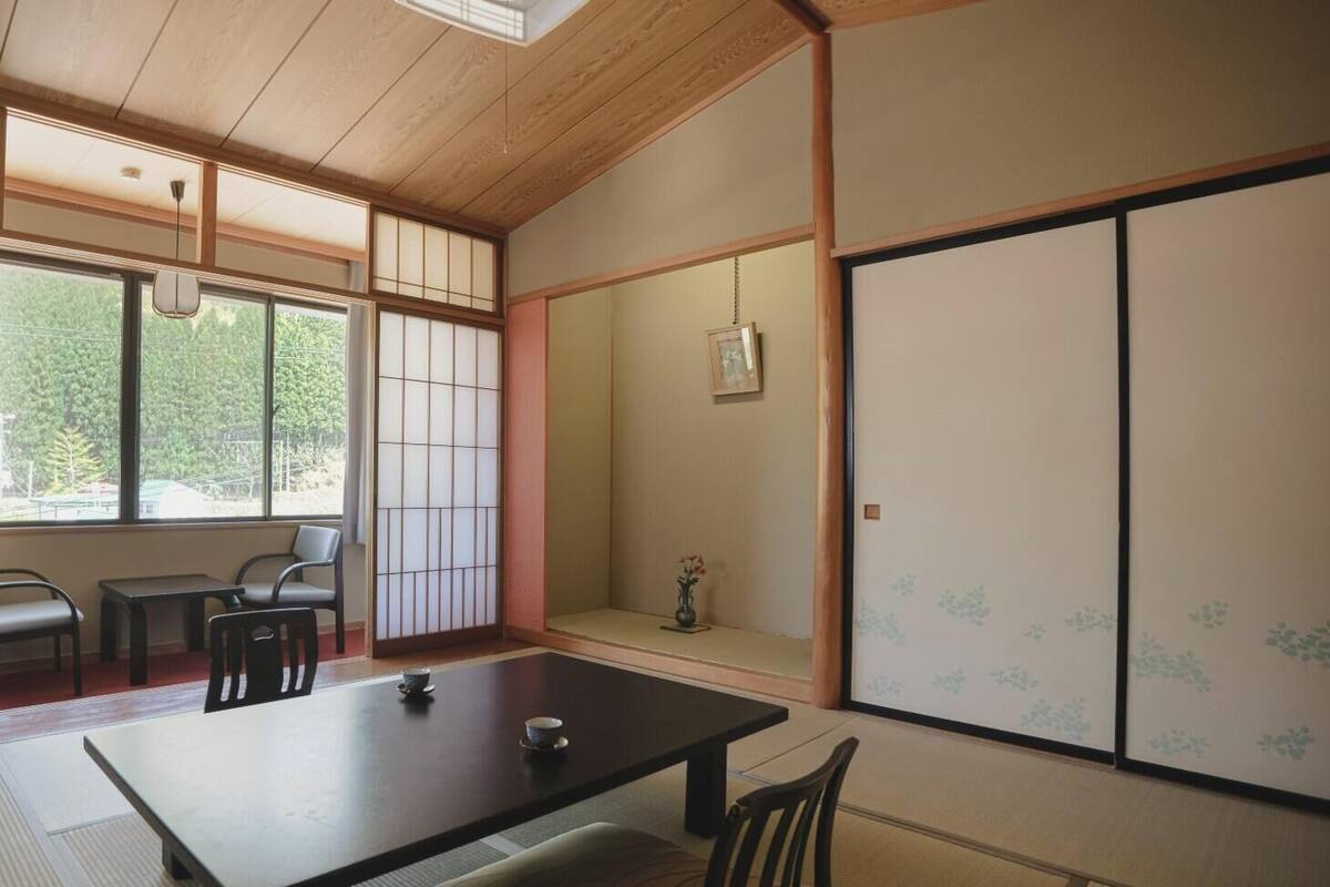 A traditional Japanese room with tatami flooring, a low table with a cushion, a sliding door with floral patterns, and a view of greenery outside.