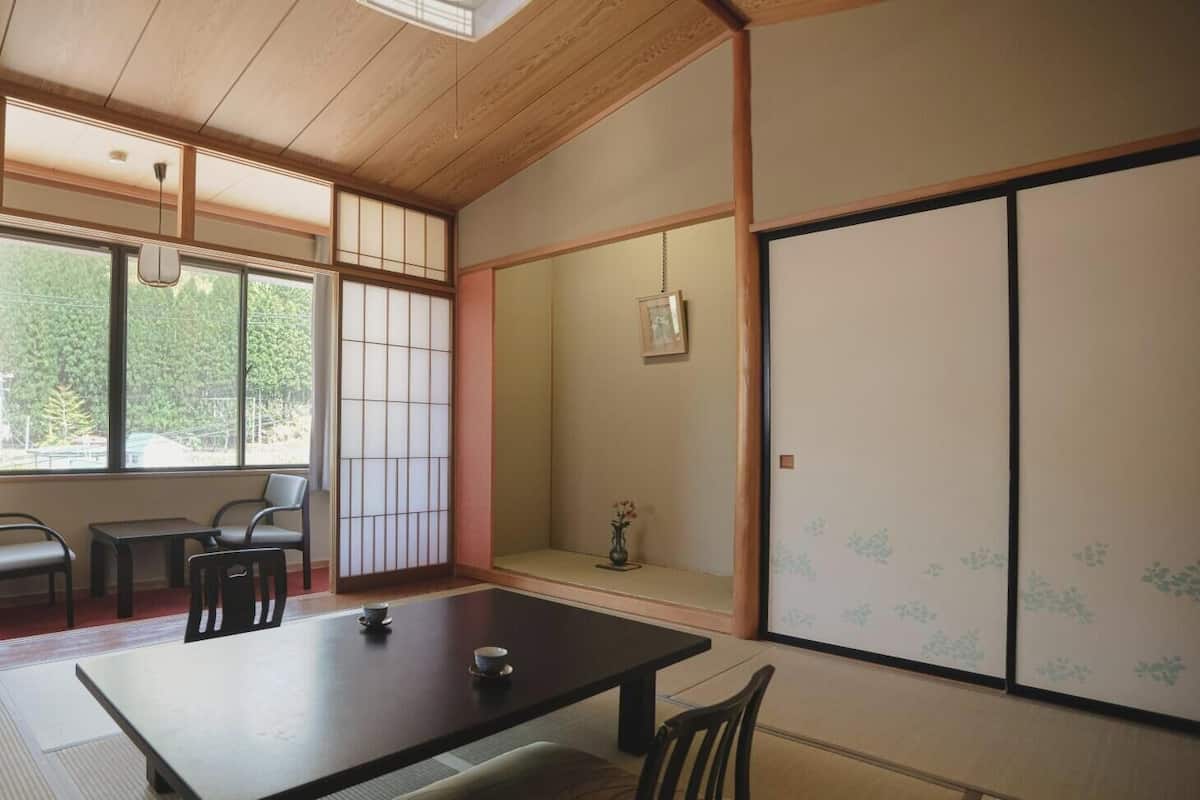 Una habitación japonesa tradicional con suelo de tatami, una mesa baja con cojín, una puerta corredera con motivos florales y vistas a la vegetación exterior.