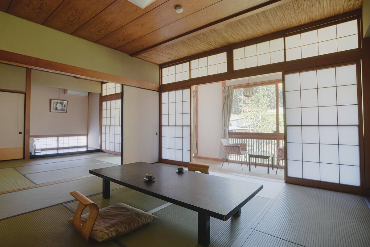 A traditional Japanese room with tatami flooring, a low table, sliding doors, and a view through large windows.