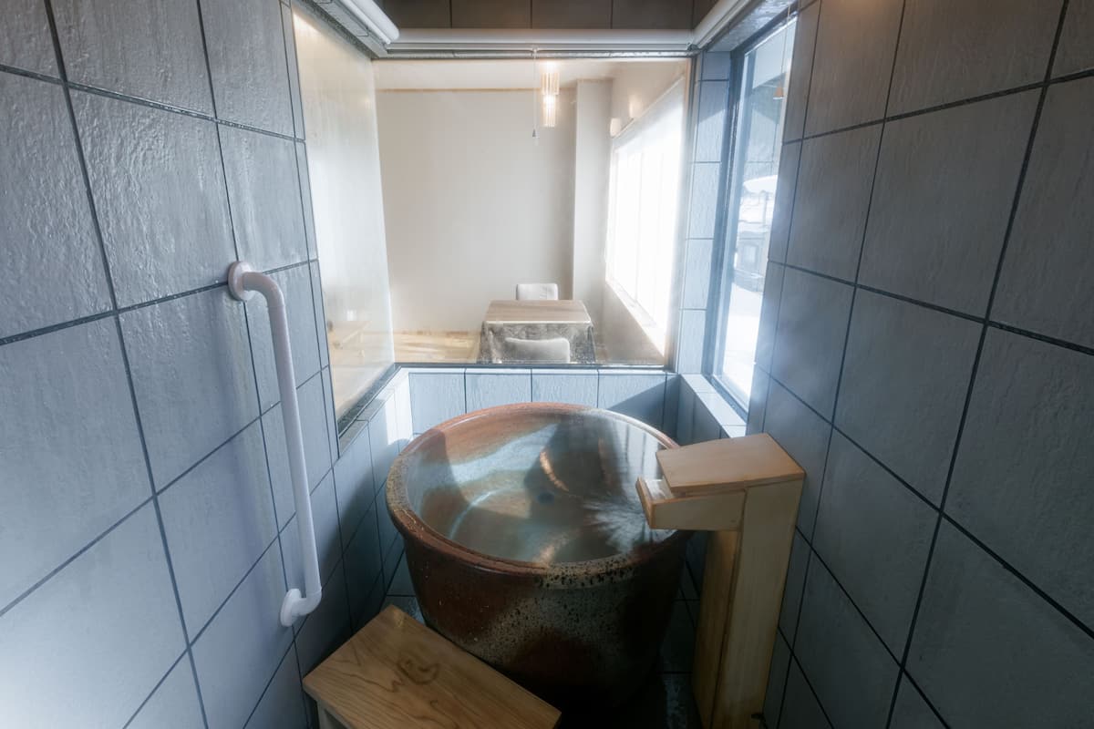A small, tiled room with a large, round, brown bathtub, a wooden bench, and a window.