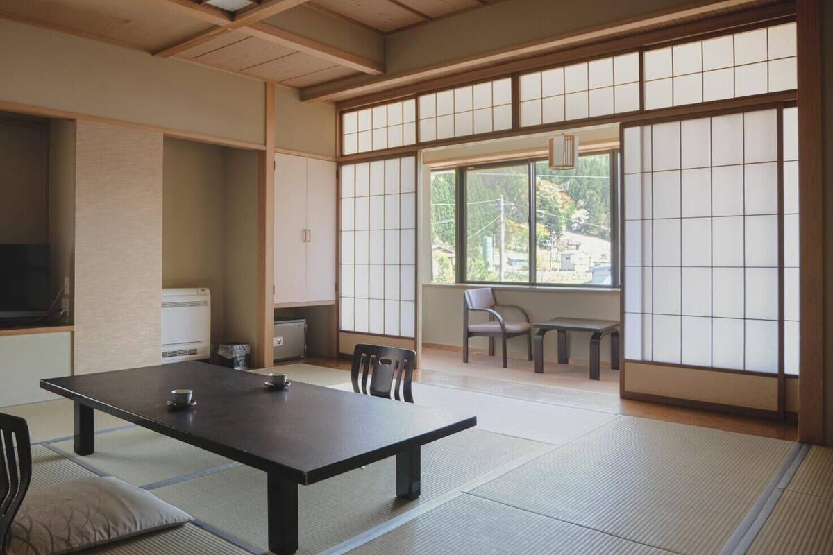 A traditional Japanese room with tatami flooring, a low table with cushions, a sliding door with shoji screens, and a view of greenery outside.