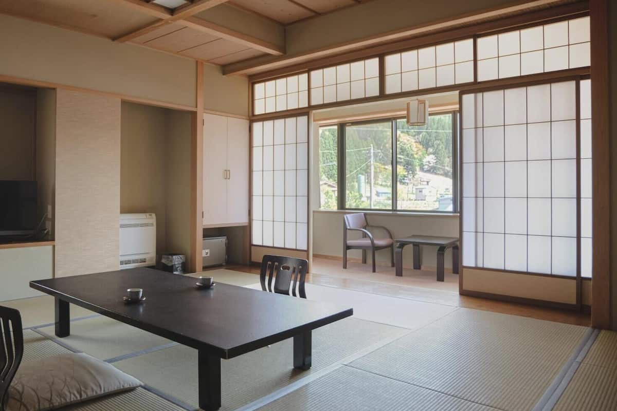Una habitación japonesa tradicional con suelo de tatami, una mesa baja con cojines, una puerta corredera con paneles shoji y vistas a zonas verdes exteriores.