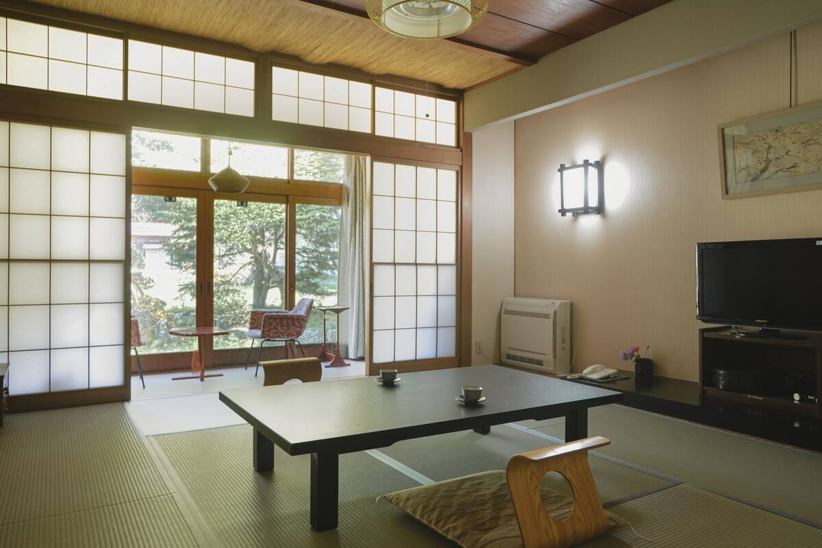 A traditional Japanese room with tatami flooring, a low table, a television, and a sliding door with a view of greenery outside.