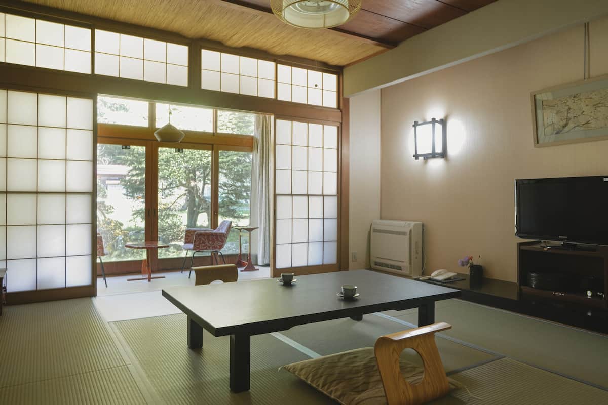 Una habitación japonesa tradicional con suelo de tatami, una mesa baja, un televisor y una puerta corredera con vistas a zonas verdes.