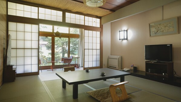 Una habitación japonesa tradicional con suelo de tatami, una mesa baja, un televisor y una puerta corredera con vistas a zonas verdes.