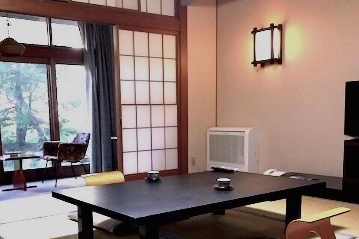 A room with a tatami mat floor, a low table with chairs, a television, and a window with curtains.