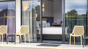 Et moderne hotelværelse med en stor skydedør, to fletstole, en seng med hvidt sengetøj og et lille natbord.