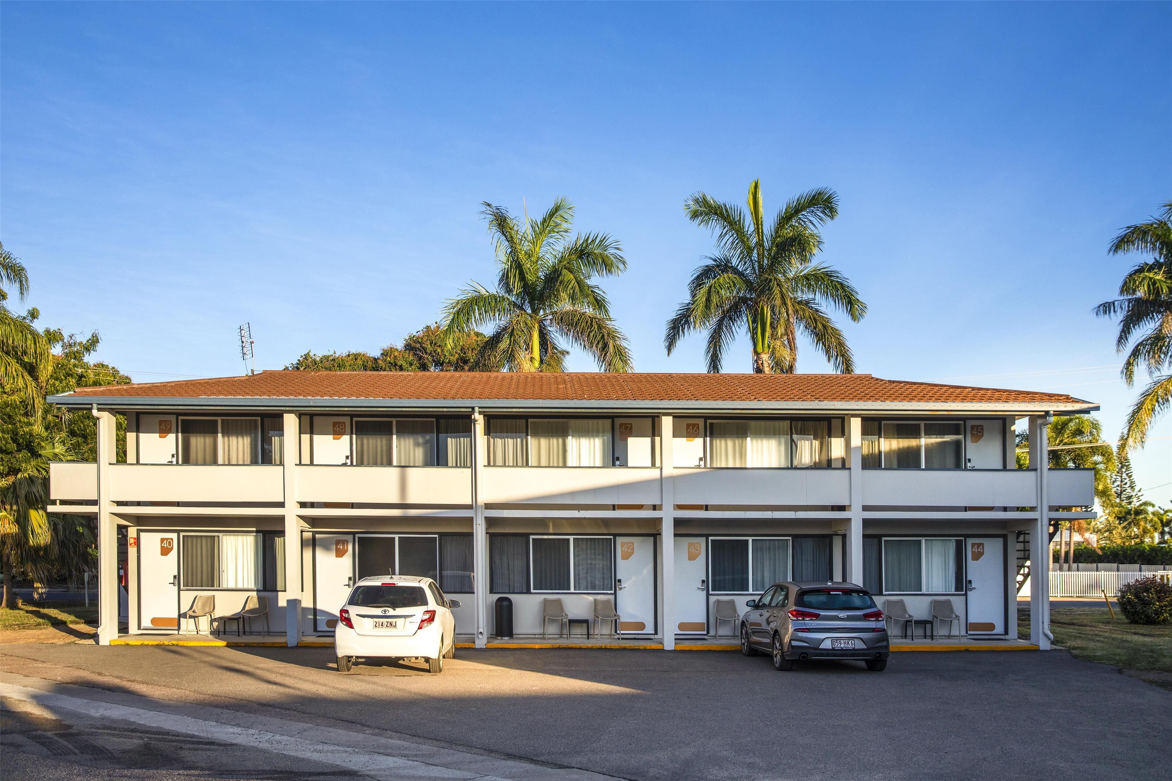 A two-story motel with a red-tiled roof, multiple rooms, and a parking area with two cars.