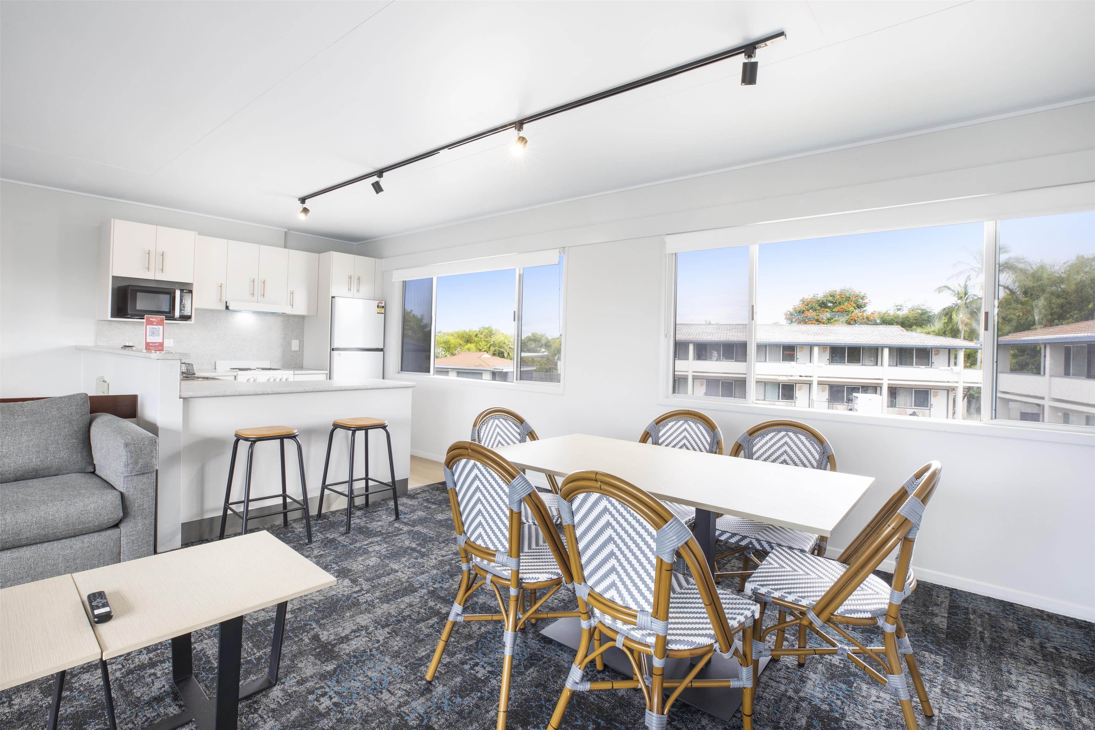 A modern kitchen with white cabinets and a microwave, a dining area with a white table and wicker chairs, and large windows offering a view of buildings and trees.
