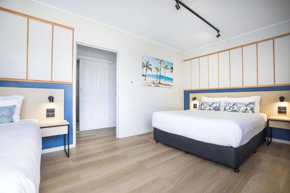 A hotel room with a large bed, bedside tables, a nightstand, a door, and a wall-mounted light fixture.