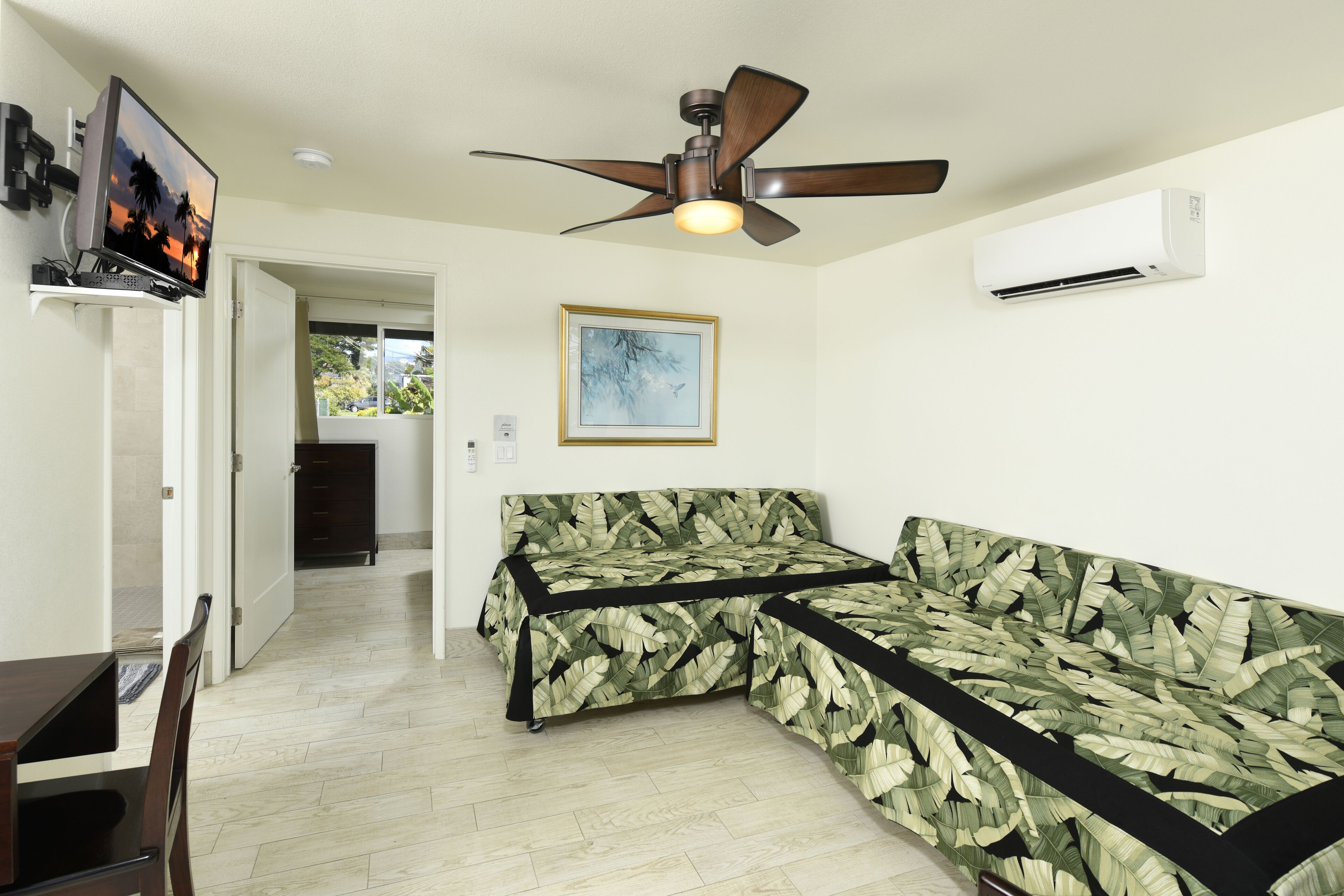 A room with a ceiling fan, an air conditioning unit, a television on a stand, a framed picture on the wall, and a bed with a leaf-patterned cover.