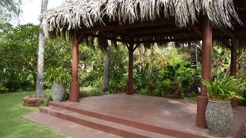 A covered outdoor area with steps leading up to a thatched roof structure, surrounded by lush greenery and potted plants.