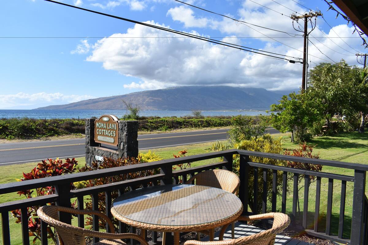 A view from a porch with wicker furniture, a sign for 'Nona Lani Cottages', and a road leading to a mountainous landscape.