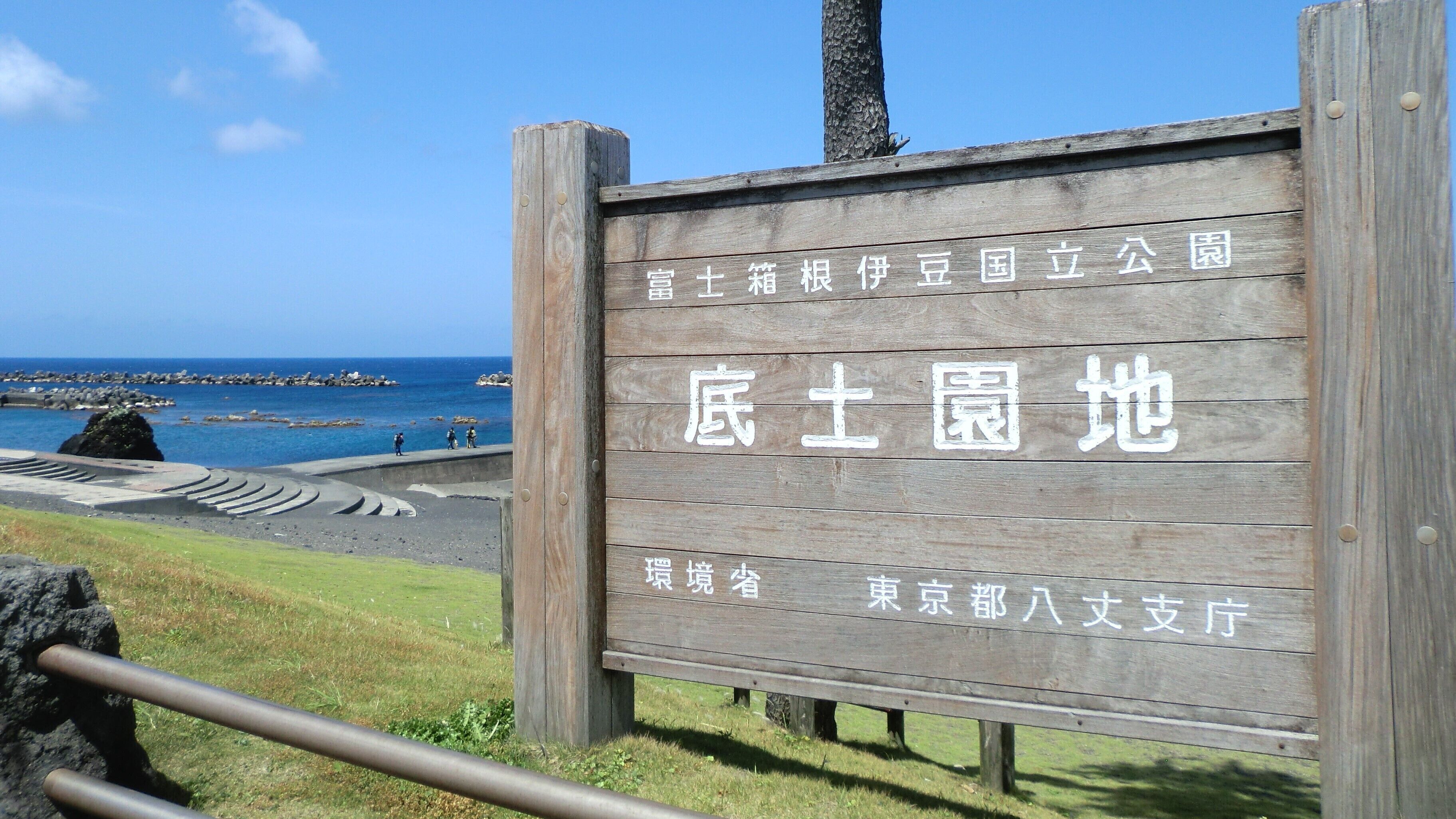 Un panneau en bois avec du texte en japonais, une vue sur la côte avec des rochers et la mer, et une zone herbeuse.