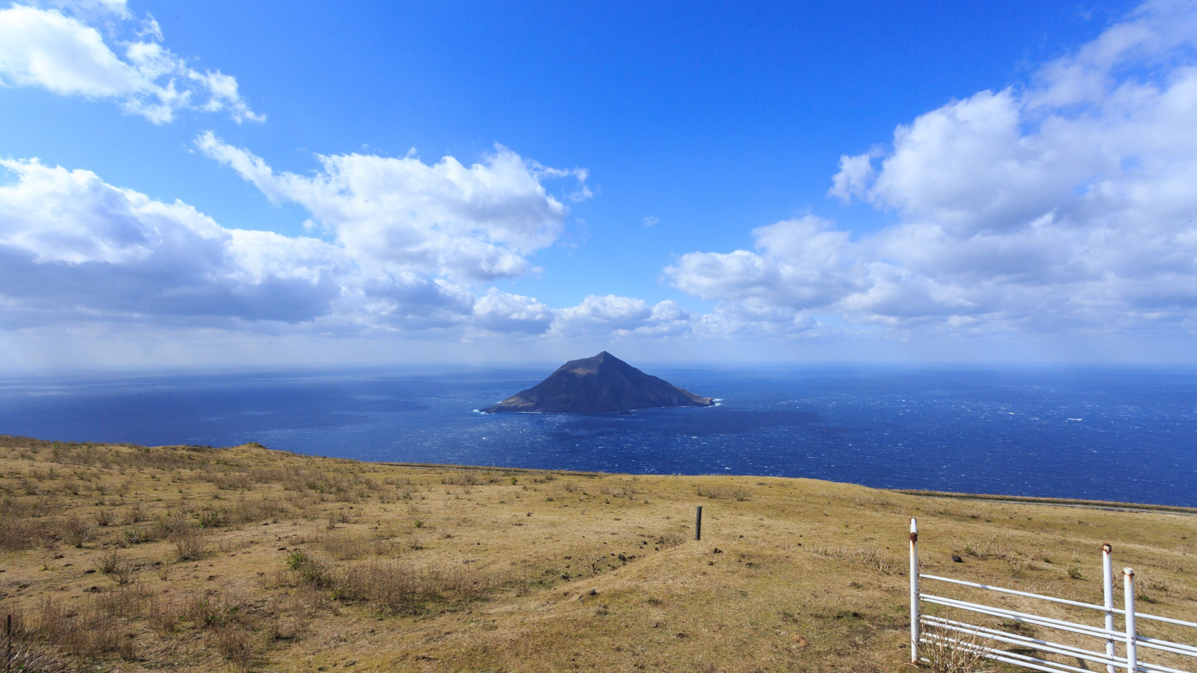 A coastal landscape with a grassy foreground, a metal fence, and a distant island with a mountain peak.