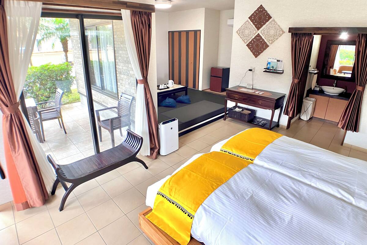 A hotel room with a bed, a desk, a chair, a small table, a sink, and a balcony with chairs.