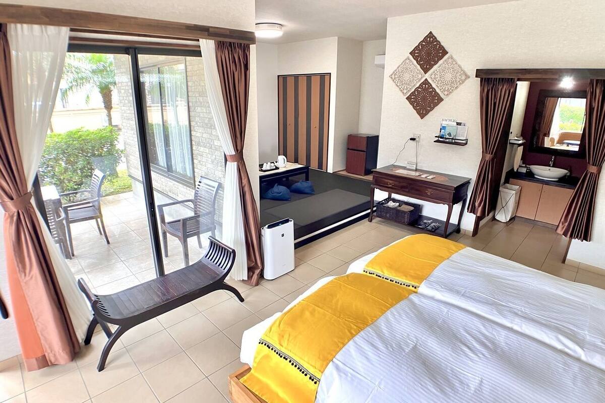 A hotel room with a bed, a desk, a chair, a small table, a wardrobe, and a sliding glass door leading to an outdoor area.