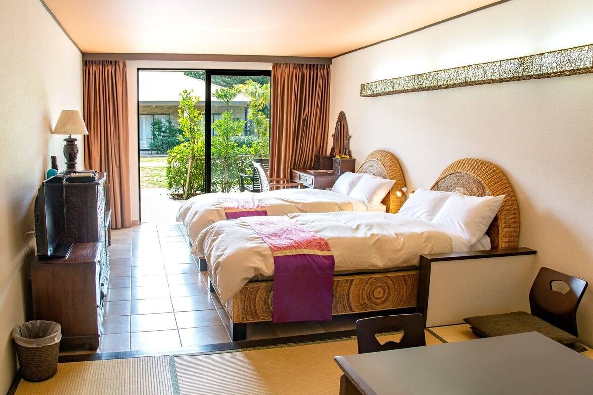 A hotel room with two beds, a desk, a chair, a TV, and a view of the outdoors.