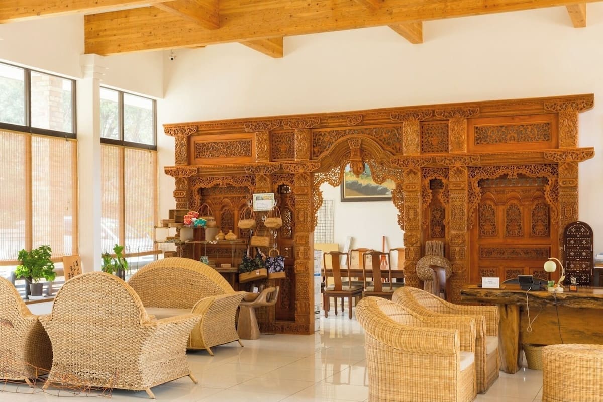 A spacious room with high ceilings and exposed wooden beams, furnished with wicker chairs and a wooden cabinet with intricate carvings.